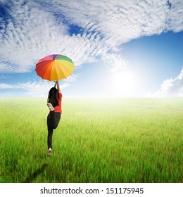 Red woman holding multicolored umbrella in green grass field and cloud sky 