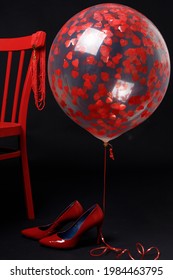 Red Women's High-heeled Shoes Near A Red Chair On A Black Background. Above Them Is A Transparent Balloon With Red Hearts.