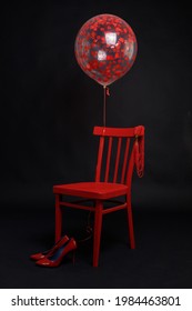 Red Women's High-heeled Shoes Near A Red Chair On A Black Background. Above Them Is A Transparent Balloon With Red Hearts.