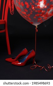 Red Women's High-heeled Shoes Near A Red Chair On A Black Background. Above Them Is A Transparent Balloon With Red Hearts.