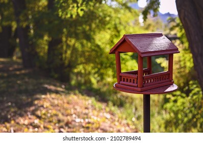Red wooden bird feeder (birdhouse) with a roof in a city park. Sunny day, autumn. The feeder stands on a metal stand on the ground. Free space for text.