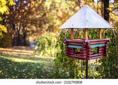 Red wooden bird feeder (birdhouse) with a metal roof in a city park. Sunny day, autumn. The feeder stands on a metal stand on the ground. Free space for text.