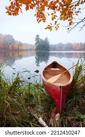 Red Wooden Canoe On Calm Lake With Trees In Fall Color And Maple Branches Above During Autumn