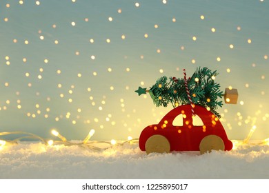 Red Wooden Car Carrying A Christmas Tree Over Snow In Front Of Blue Background And Golden Garland Lights