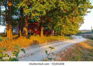 Red Wooden Cottage In Oak Forest. Beautiful Sunny Evening In Autumn, Last Sun Beams Of The Day. Autumn Season With Yellow Leaves And Yellow Grass. Brick Pathway. Big Rocks Near Road.