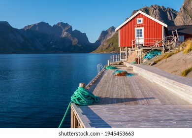 Red Wooden Fisherman's House In Reine, Lofoten, Norway. Sunset Of A Sunny Day In The Village By The Sea. Wooden Pier And Beautiful Small Wooden House On The Shore. Sunset In The Fjord. North.