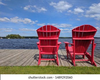 Red wooden Muskoka Adirondack chairs at a pier in front of the lake, beautiful sunny day, blue sky with white clouds. Muskoka, Ontario, Canada. Space for copy.