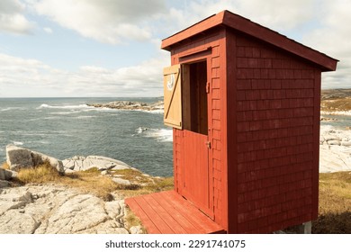 Red wooden outhouse,  closeup, with 2 part door, a slanted roof and shingle siding, on a rocky shore, looking over the ocean. The upper half of the door is open with a moon, bathroom, sign - Powered by Shutterstock