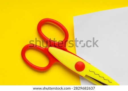 Red and yellow craft scissors with a zigzag blade cutting white paper on a bright yellow background