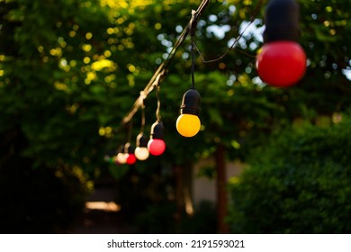 Red And Yellow Light Bulbs Garland Over The Path In The Garden. Garden In The Backyard Of The House. Landscape Design.