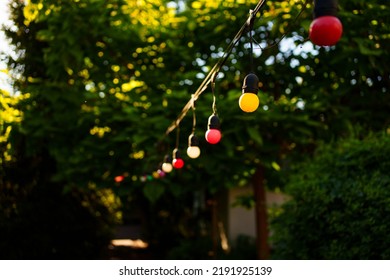 Red And Yellow Light Bulbs Garland Over The Path In The Garden. Garden In The Backyard Of The House. Landscape Design.