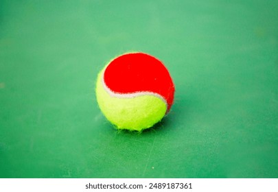 A red and yellow tennis ball sits on a green court. Ideal for highlighting tennis gear for beginners or junior players. - Powered by Shutterstock