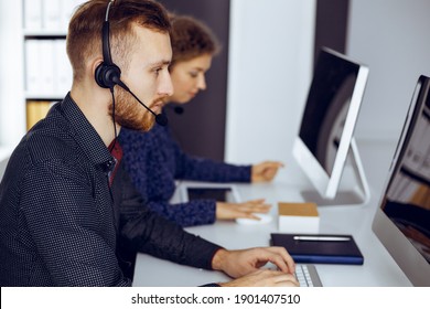Red-bearded businessman talking by headset near his female colleague while sitting in modern sunny office. Diverse people group in call center