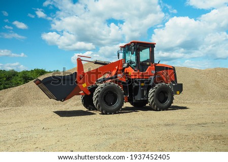 A red-black front-end loader with small wheels against the background of a large pile of stone sand and a blue sky with white clouds. Side view.