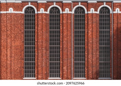 A reddish masonry wall with long gray windows. A red brick facade, white moldings and downpipes. A texture of a vintage building wall with elongated rounded vertical crevice windows and white boiserie