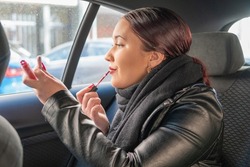 Reddish-haired Teenage Girl With Black Jacket Inside A Vehicle With Lipstick While Looking At The Mobile. Young Woman Holding A Lipstick And Looking On The Mobile To Put On Makeup And Be Perfect.