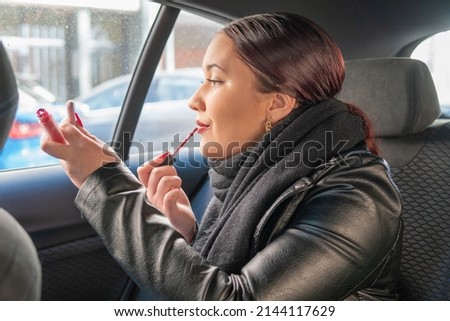 Reddish-haired teenage girl with black jacket inside a vehicle with lipstick while looking at the mobile. Young woman holding a lipstick and looking on the mobile to put on makeup and be perfect.