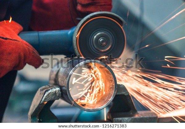 A red-gloved mechanic cleans a welded seam on a\
section of a steel pipe with the help of a grinding machine in the\
metal workshop