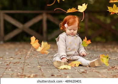 Red-haired Baby Girl On The Background Of Nature And Sitting In The Fall Leaves