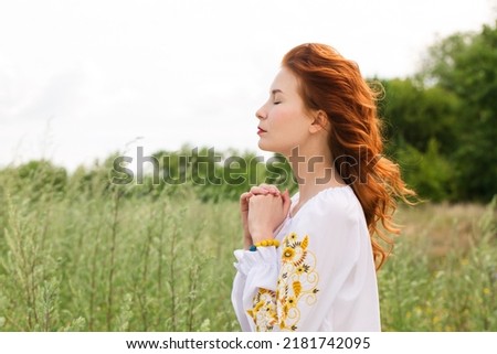 Red-haired, beautiful, young woman in a Ukrainian embroidered shirt prays for Ukraine in the field
