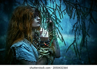 Red-haired Druid Woman With A Mask On Eyes Stands In A Gloomy Forest Among The Tree Branches In The Rain Trying To Awaken Her Spiritual Vision. Paganism. Halloween. 