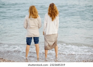 Red-haired elderly mother and her grown-up daughter are walking along the seashore together. Happy meeting of a mother and her grown-up child. Family relations between adult children and older parents - Powered by Shutterstock