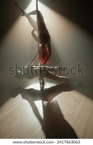 Red-haired female performer on pylon reaching hand to male partner standing in planche on stage floor during pole dance in dim spotlight