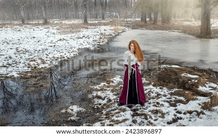 A red-haired girl in an ancient dress in a meadow among the spring melting snow.