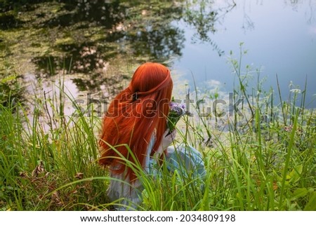 Redhaired girl with flowers in her hands sits in the grass near a picturesque lake. View from the back. Long hair. Fairy tale.