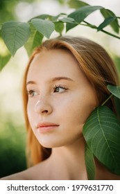 Red-haired girl with freckles close-up stands near the leaves on the background of greenery without makeup looks away