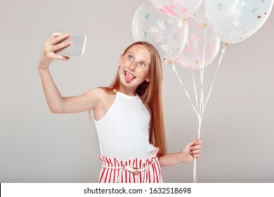 Red-haired girl with freckles standing isolated on grey background inclusive beauty holding balloons taking selfie photo on smarthpone showing tongue smiling silly - Powered by Shutterstock