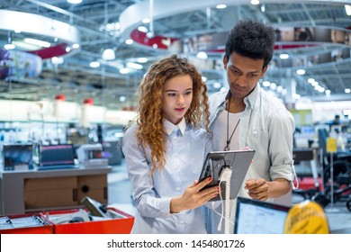 Red-haired Girl With Glasses With An African Guy In An Electronics Store Standing In Front Of The Stand Choosing A Tablet Computer To Buy