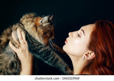 Red-haired Girl Holds Fluffy Cat On Black Background. Pretty Woman With Folded Lips Wants To Kiss Pussycat. Love For Pets.