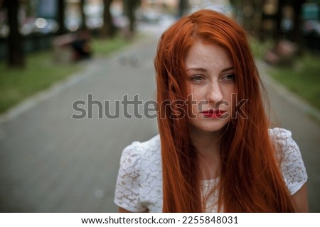 a red-haired girl in a light skirt; a very cheerful girl with long red hair; lips painted with red lipstick; sits on a bench and has a good mood; poses for a photographer; street photo session 