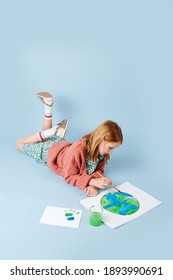 Red-haired Girl Lying On The Floor Draws A Green And Blue Planet. Studio Shot