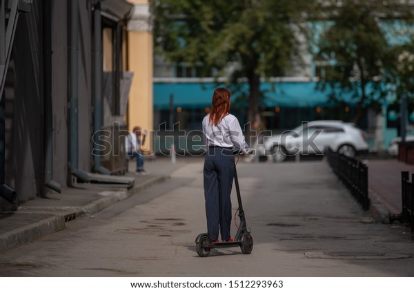 A red-haired girl in a white shirt drives an\
electric scooter along the wall. A business woman in a trouser suit\
and red high heels rides around the city in a modern car. dress\
code in the office.