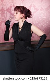 red-haired lass wearing black gown and long black gloves is standing and having alcohol