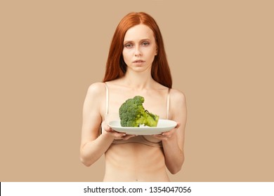 Red-haired neutral emotional neutral woman in uderwear looking at camera, holding plate with raw broccoli over beige background. Concept of weight loss, diet, eating disorders, anorexia.