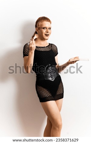 A red-haired non-binary person is posing in extravagant clothes on a white background happily. The man wears a brush and a makeup palette. Concept of people is not identified with any gender.