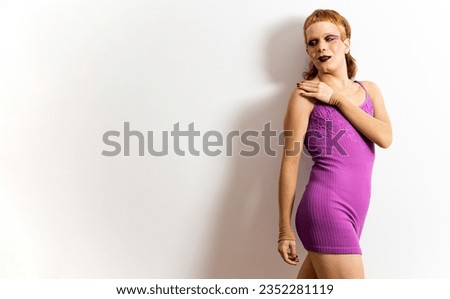 A red-haired non-binary person is posing on a white background in a purple dress while touching one shoulder and looking to the side. Concept of non-binary models. People without gender.Copyspace.