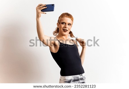 A red-haired non-binary person is posing on a white background in extravagant clothing and takes a picture with the mobile phone while pulling his hair. Concept of social networks. Relate socially.