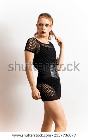 A red-haired non-binary person is posing on a white background while pulling his hair. The man is wearing extravagant clothes with a mesh and corset.Concept of people is not identified with any gender