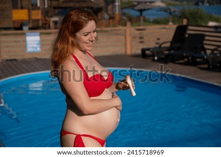 A red-haired pregnant woman sunbathes by the pool. The expectant mother applies sunscreen to her tummy.