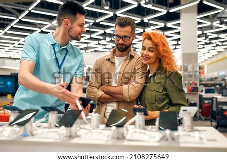 Red-haired sweet woman, along with her boyfriend, chooses new smartphone in store of household appliances, electronics and gadgets, getting help from store consultant.