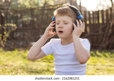 A red-haired teenager boy with headphones in a white T-shirt listening to music on a mobile tablet and relaxing on the grass in the fresh air in nature - Powered by Shutterstock