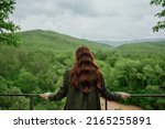 a red-haired woman with beautiful, well-groomed, long hair stands with her back to the camera and enjoys the view of the forest. High quality photo