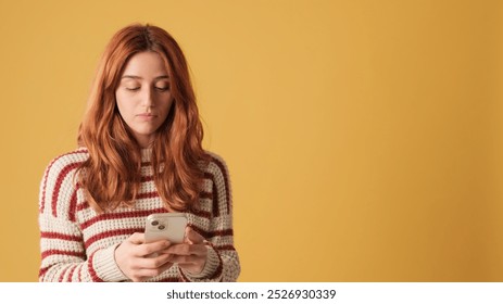 Red-haired woman dressed in sweater, texting on mobile phone on yellow background. Beautiful girl poses in studio with blank space on yellow wall for your promotion - Powered by Shutterstock