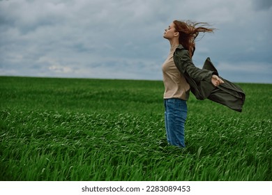a red-haired woman in a long coat stands in a green field and the wind blows her hair
