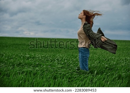a red-haired woman in a long coat stands in a green field and the wind blows her hair