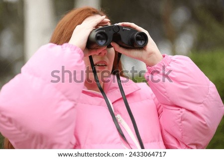 Red-haired woman, with a pink coat, observes the birds in the trees of a park with binoculars